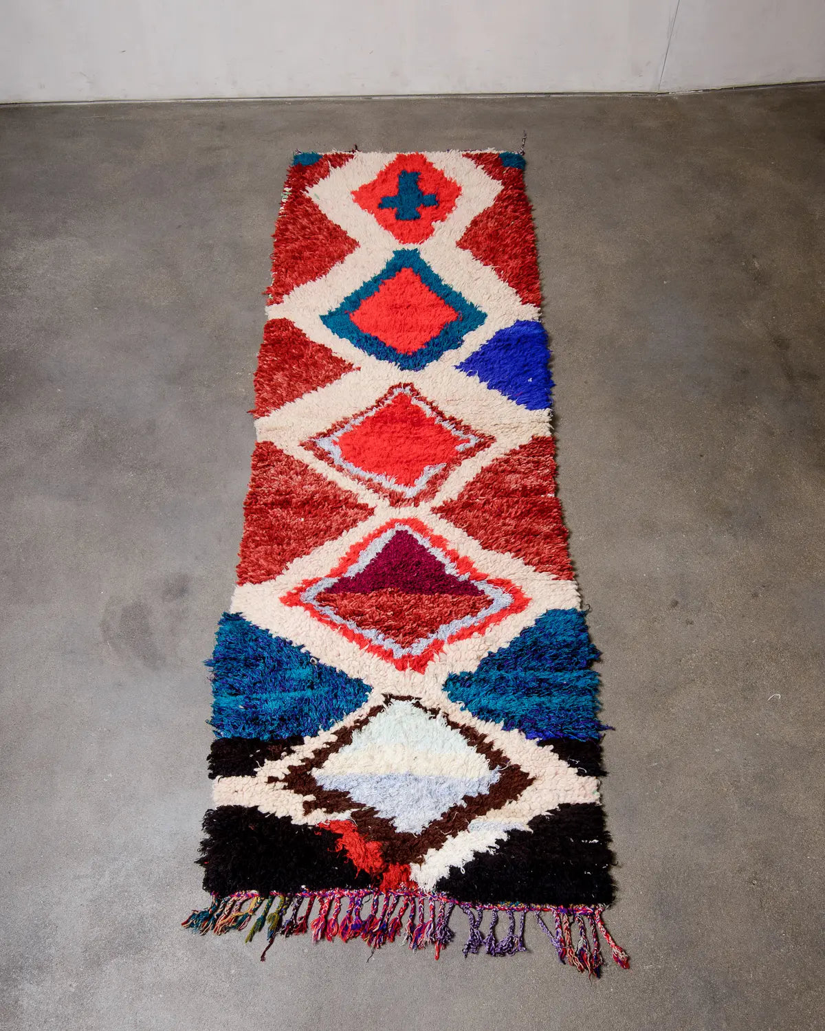 Runner rug with design in predominantly red, cream and blue laying on grey concrete floor.